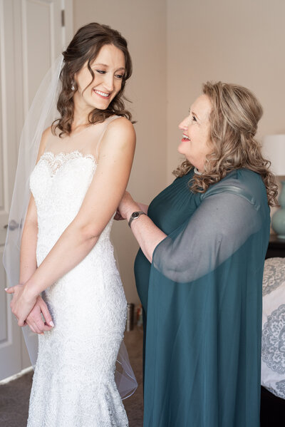 Mother of bride fastens the back of her daughters wedding gown as she smiles