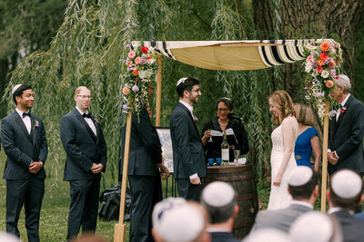 Bride and groom stand under a floral chuppah during an outdoor wedding ceremony at Barnwood Events in Madison, Wisconsin.