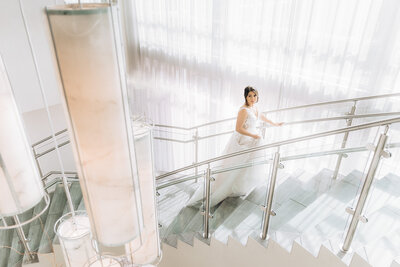 An image of a bride wearing a flowing white dress as she walks up a modern glass and marble staircase with large floor to ceiling windows behind her and a chandelier in the foreground at the Embassy Suites Hotel in Greenville SC, taken by Zack Bradley Photography