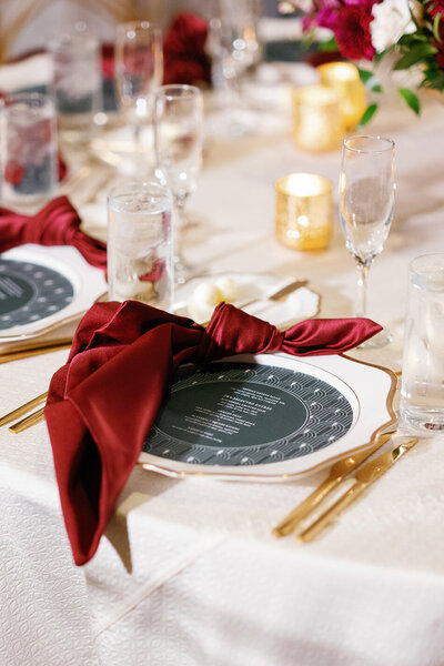 Festive place setting at the Don Cesar Hotel in St. Pete Beach, FL, featuring white linens, gold edge scalloped chargers, scarlet red knotted napkins, and round green wedding menus nestled inside the chargers.