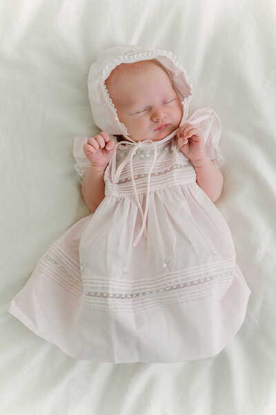 Baby laying on a white sheet weraring a heirloom newborn gown and bonnet.