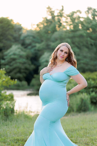 A beautiful mom-to-be posing in a blue dress in front of a lake.