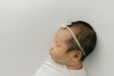 Profile image of newborn baby girl with a bow on her head in all white studio by New Jersey baby photographer