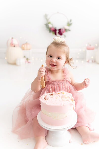 A young toddler girl in a pink tule dress sits in a Greater Atlanta Motherhood & Family Photographer studio smashing a pink cake