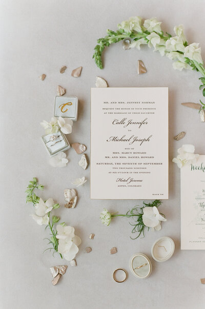 Flat-lay of white wedding invitation with cursive writing, green stems, white flowers and pink flower-petals, and wedding rings