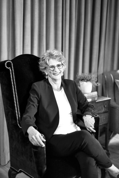 A black and white photograph of a smiling woman, Amy Posner, seated in an elegant chair.