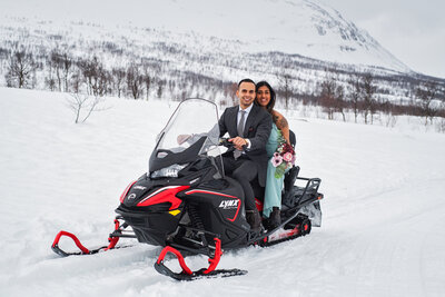 A happy couple is celebrating their wedding day in Norway, posing on a red and black snowmobile against a winter landscape. The groom, in a smart suit, is at the helm of the snowmobile, and the bride sits behind him, embracing him and smiling broadly. She is wearing a pastel green dress and holds a bouquet of light and dark pink flowers. They are surrounded by a vast expanse of snow with a backdrop of mountains, capturing the unique spirit of a Norwegian winter wedding.