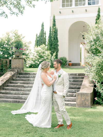Bride and groom standing together and smiling at each other outside a venue surrounded with greenery and trees