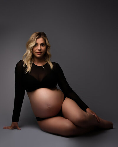 Expectant mother in her second trimester seated on the floor on a grey background wearing a black crop top with dramatic lighting to accentuate her baby bump