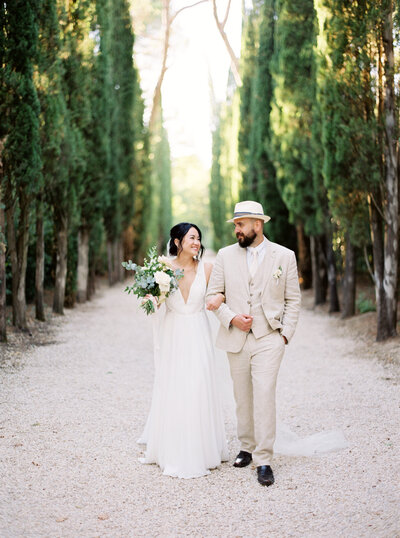 Bride smiles at her groom while holding bouquet as they walk down tree lined path