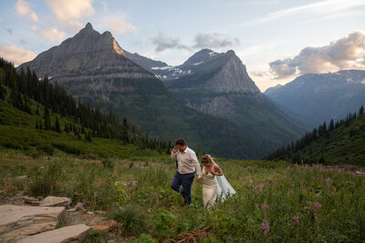 A bride and groom walk hand in hand along a path surrounded by wildflowers in Glacier national Park, Montana.