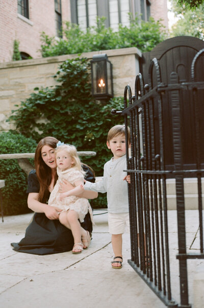 Film photograph of mother holding her daughter while her son grips a garden iron fence in downtown Chicago, capturing authentic, emotional, and intimate family moments in a relaxed documentary style.