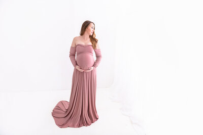 A mother to be in a pink maternity gown looks down her shoulder and holds her bump while standing in the studio of a Greater Atlanta Motherhood & Family Photographer