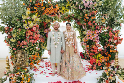 Bride and groom stand together under a vibrant floral arch at their Indian fusion wedding, surrounded by an array of multicoloured flowers.