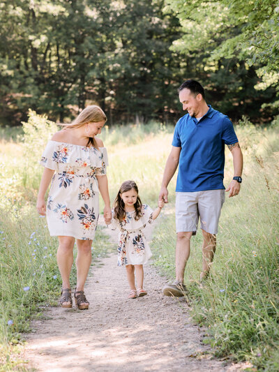 mom and dad holding hands with daughter walking in park for family photos