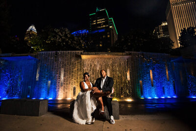 Portrait-of-black-couple-who-just-eloped-sitting-in-Romare-Bearden-Park-in-front-of-the-blue-and-yellow-waterfall-with-the-Charlotte-skyline-in-the-background