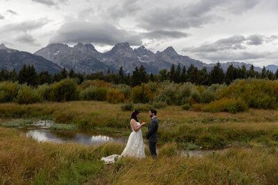 A bride reads her vows to her groom as they stand in a grassy meadow with the Grand Teton mountains behind them.