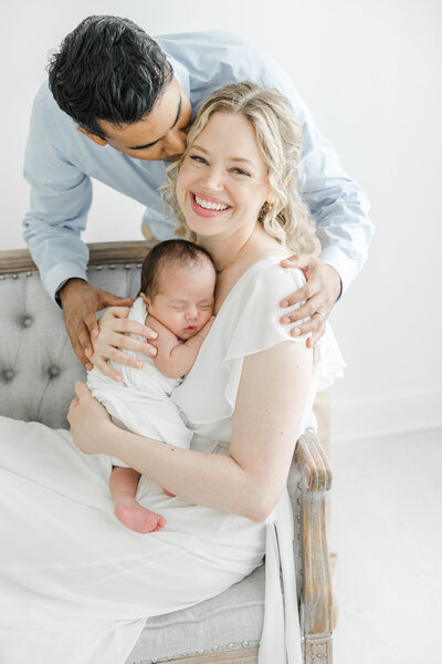 New mom smiles while holding her newborn baby while her husband kisses her cheek during newborn portrait session