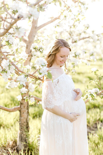 Expecting mother stands among flowering tree in the sunshine for Minneapolis maternity photoshoot.