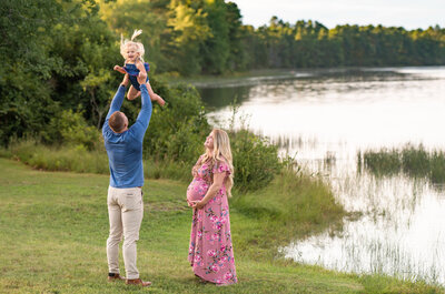 A dad wearing a blue shirt, tossing his toddler daughter in the air, while his pregnant wife, wearing a pink floral dress watches and smiles