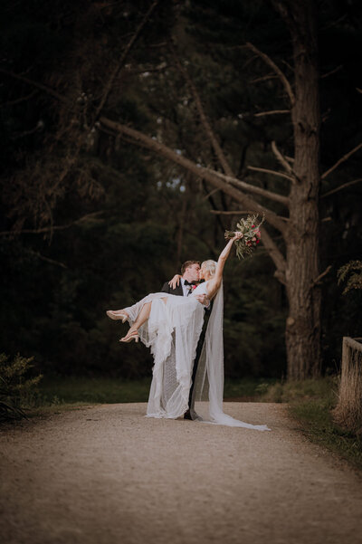 Bride & Groom on a forest path, he is carrying her while they kiss and she holds her bouquet in the air.