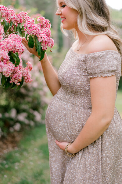 A close up of a mother holding her belly while smelling pink flowers in a brown and white dress.