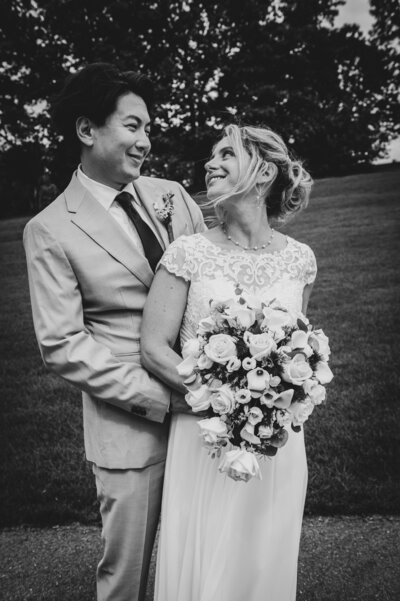 A joyful bride and groom standing together on their wedding day, holding a bouquet and smiling at each other during an intimate New England celebration. Photographed by Sarah Ferrazzani Photography.