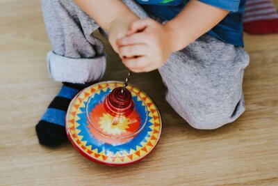 A child is playing with a colorful spinning top on a wooden floor, gripping the handle to set it in motion while wearing cozy grey pants and striped socks.