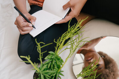 Black woman sitting by a plant writing in a notebook