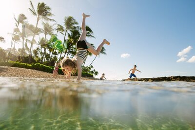 A girl does a handstand in the water as children play in the ocean behind her.