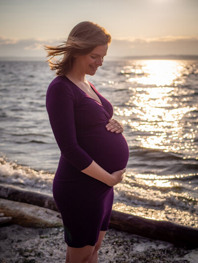 A pregnant woman stand at the shoreline at Carkeek Park, WA looking down at her pregnant belly.