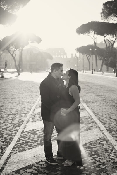 A b/w photo of a pregnant woman and her husband with hands on her belly kissing in front of the Colosseum. Taken by Rome Photographer, Tricia Anne Photography.