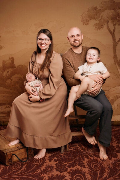 A family sits together barefoot in front of a brown-toned backdrop—a woman holding a baby and a man with a young child, captured by a photographer in Bemidji, Minnesota.