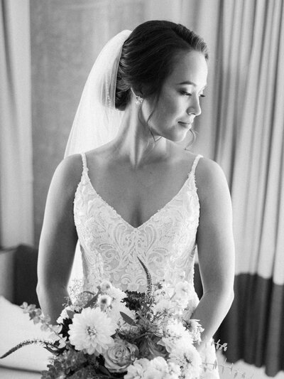 A black and white image of a bride holding her bouquet and gazing over her shoulder as window light6 beautifully lights the contours of her face