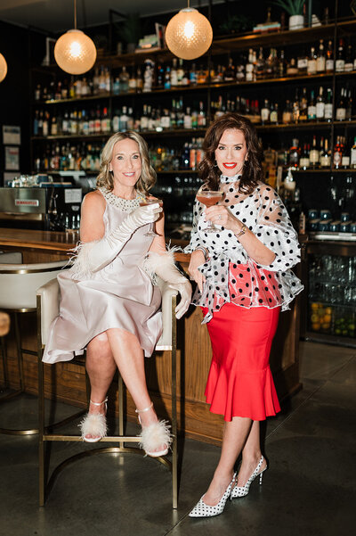 Two wedding planners enjoy cocktails at a stylish bar, showcasing the vibrant and personable approach of Socially Significant Events to event planning