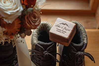 The image captures a beautifully arranged wedding detail with a pair of fur-lined hiking boots and a wedding bouquet. The bouquet, rich with white, cream, and burnt orange flowers, drapes elegantly over the edge of one boot. Resting on the other boot is a personalized wooden ring box, engraved with "Adam & Jessica, Tromsø, Norway, October 2022", alongside a mountain range motif, signifying the couple's adventure and union. The warm lighting enhances the texture of the wooden box and the intricate details of the flowers, celebrating the couple's special day in a cozy, rustic setting.
