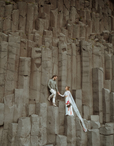 the bride and groom are standing on a basalt column, holding hands and looking at each other. the bride has on a long veil and is holding florals. the groom has on jeans and a t-shirt with his hair braided.