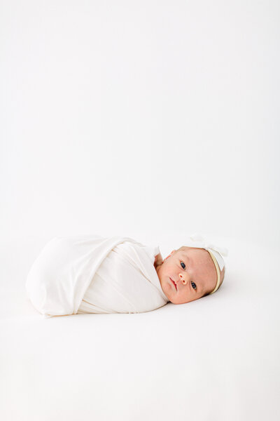 Newborn baby girl swaddled in white blanket with white bow on her head by Cincinnati Newborn Photographer