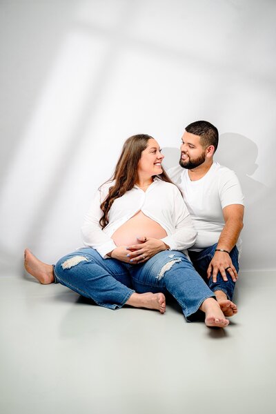 Charlotte couple sits next to each other wearing jeans and white shirts with moms expectant belly showing.