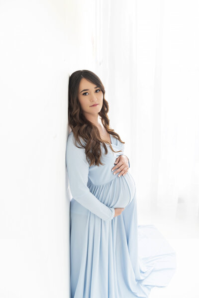 A pregnant motherleans against a wall in a studio in a blue maternity gown