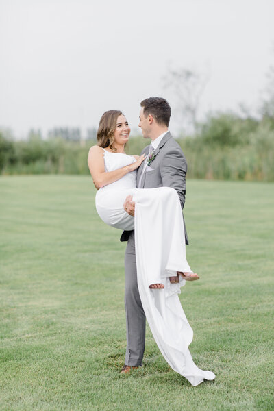 Groom carrying his bride through the grass field at Aquatopia Wedding Venue in Ottawa, Ontario. Photographed by Ottawa and Destination Wedding Photographer, Brittany Navin Photography