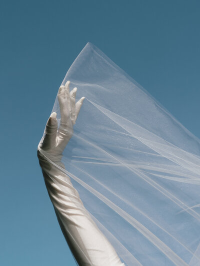 Hand wearing a white silk glove holding part of a veil against a blue sky background