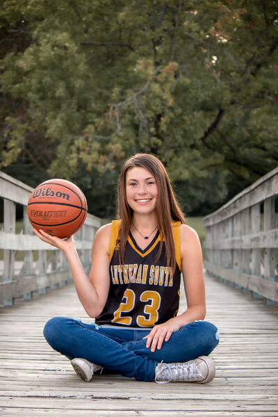 High school senior girl sitting criss cross applesauce on a bridge holding a basketball up in her right hand