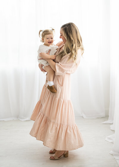 Mom in pink dress, holding her daughter while smiling at a studio in Ottawa