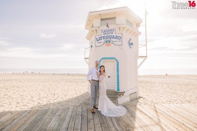 Groom to be holds his Bride from behind as they pose for engagement photos on the sands with ocean waters of Main Beach Laguna