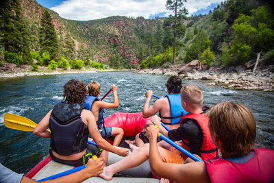 A group of people wearing life jackets are rafting down a river surrounded by lush green trees and rocky banks. They are paddling together in a red inflatable raft, with a scenic view of hills in the background.