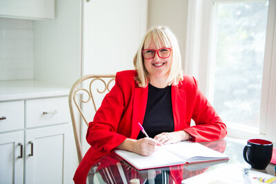 Christine DeHerrera in red glasses, red blazer sitting in a chair at a glass table with a  notebook and pen