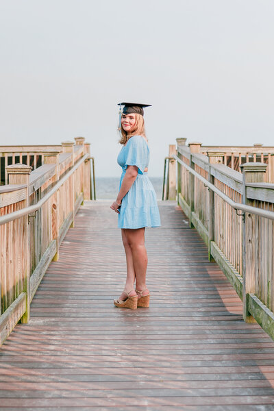 high school senior standing on dock at a beach with senior cap on a sunny day