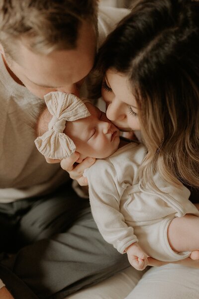 Sweet newborn baby girl wearing a beige bow is kissed by bother her parents