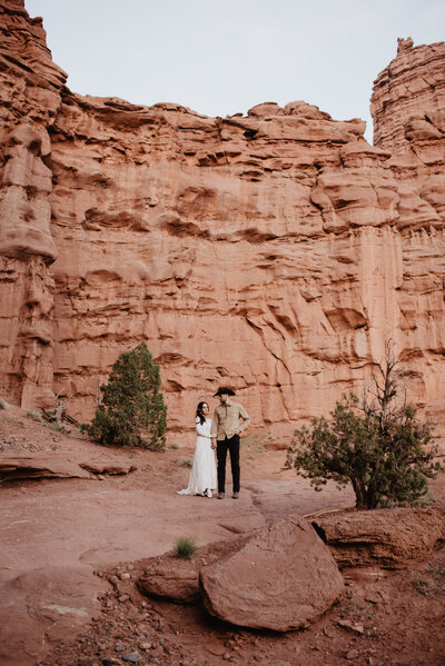 utah elopement photographer captures cowboy wedding in Moab with bride and groom standing in front of a large red rock cliff